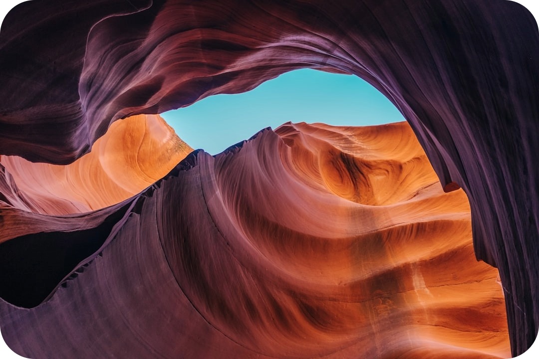 Looking up through a sandstone slot canyon, with wave-like orange and purple rock walls framing a strip of blue sky.
