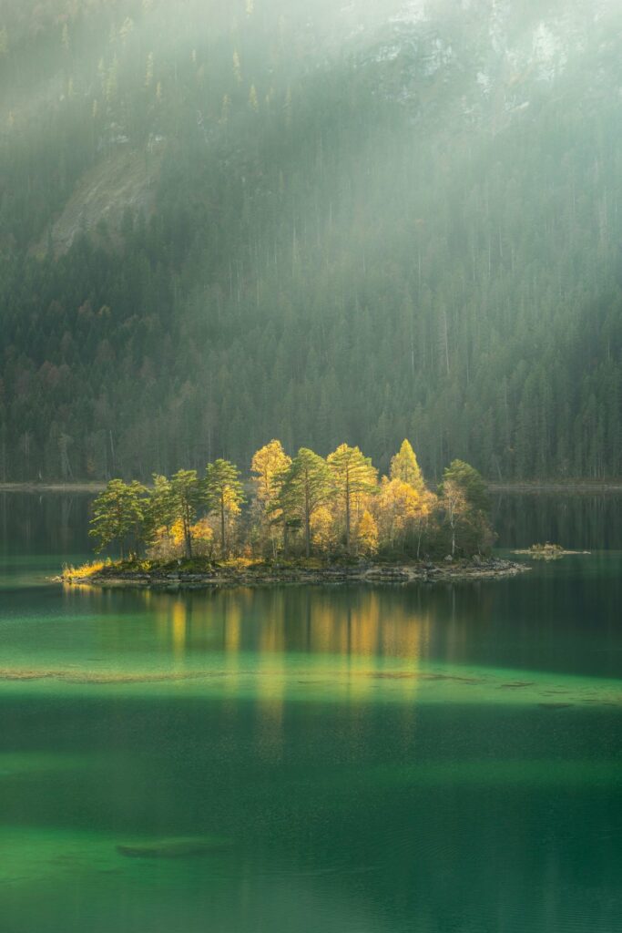 A small island of pine and golden autumn trees lit by shafts of sunlight, sitting on a calm emerald-green lake with a misty, forested mountainside rising behind it.