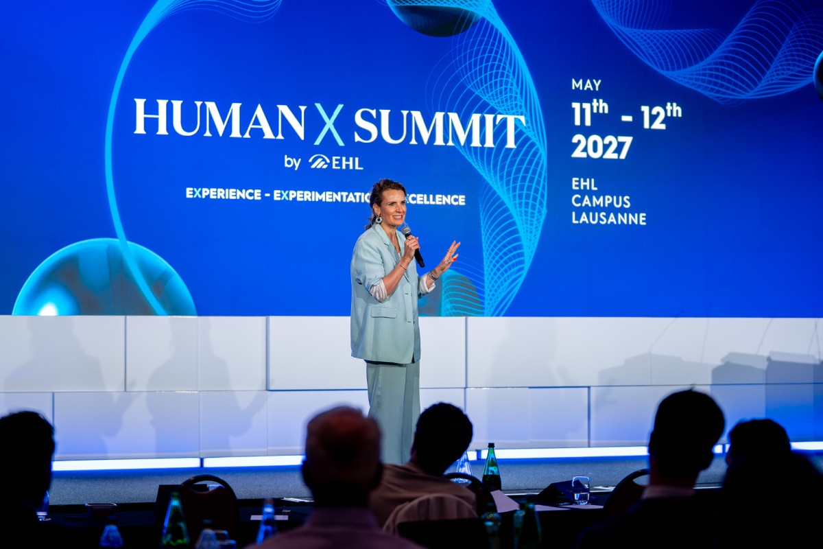 Master of ceremony in a light blue suit speaking with a microphone on stage in front of a large blue screen reading "Human X Summit by EHL, May 11th–12th 2027, EHL Campus Lausanne," with audience members seated in the foreground.