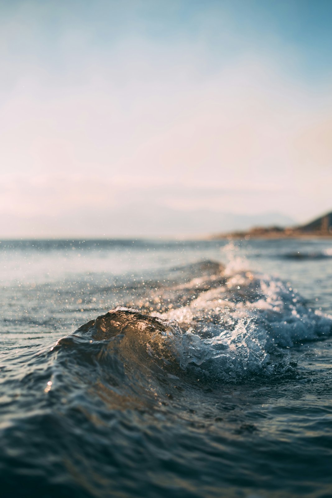 A close-up of a small ocean wave cresting and splashing at water level, with sunlit droplets scattered in the air, under a soft pastel sky with a blurred coastline in the distance.
