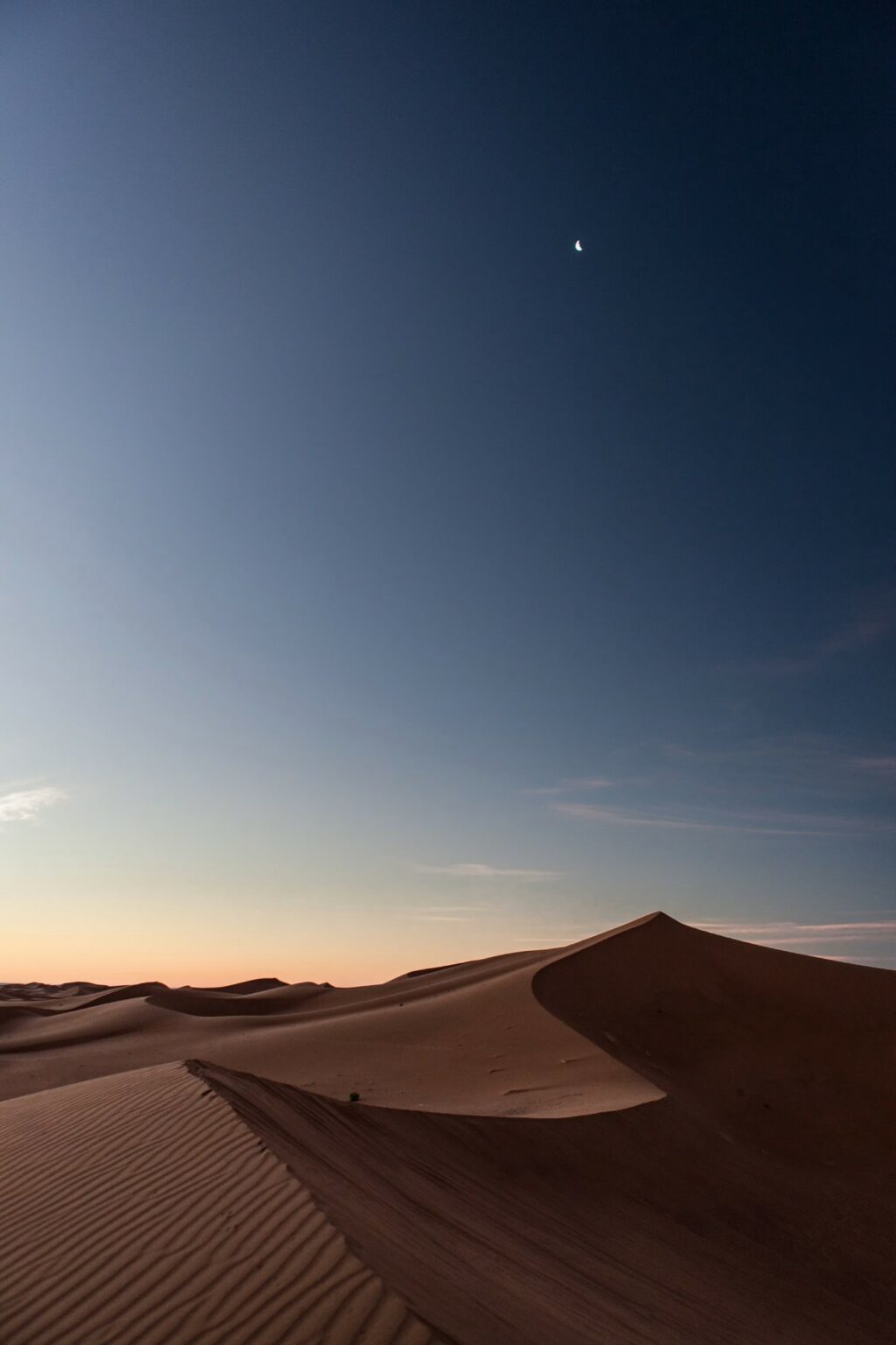 Rippled sand dunes at dusk under a deep blue sky fading to pale orange at the horizon, with a small crescent moon high overhead. A sharp dune ridge curves through the foreground, casting long shadows.