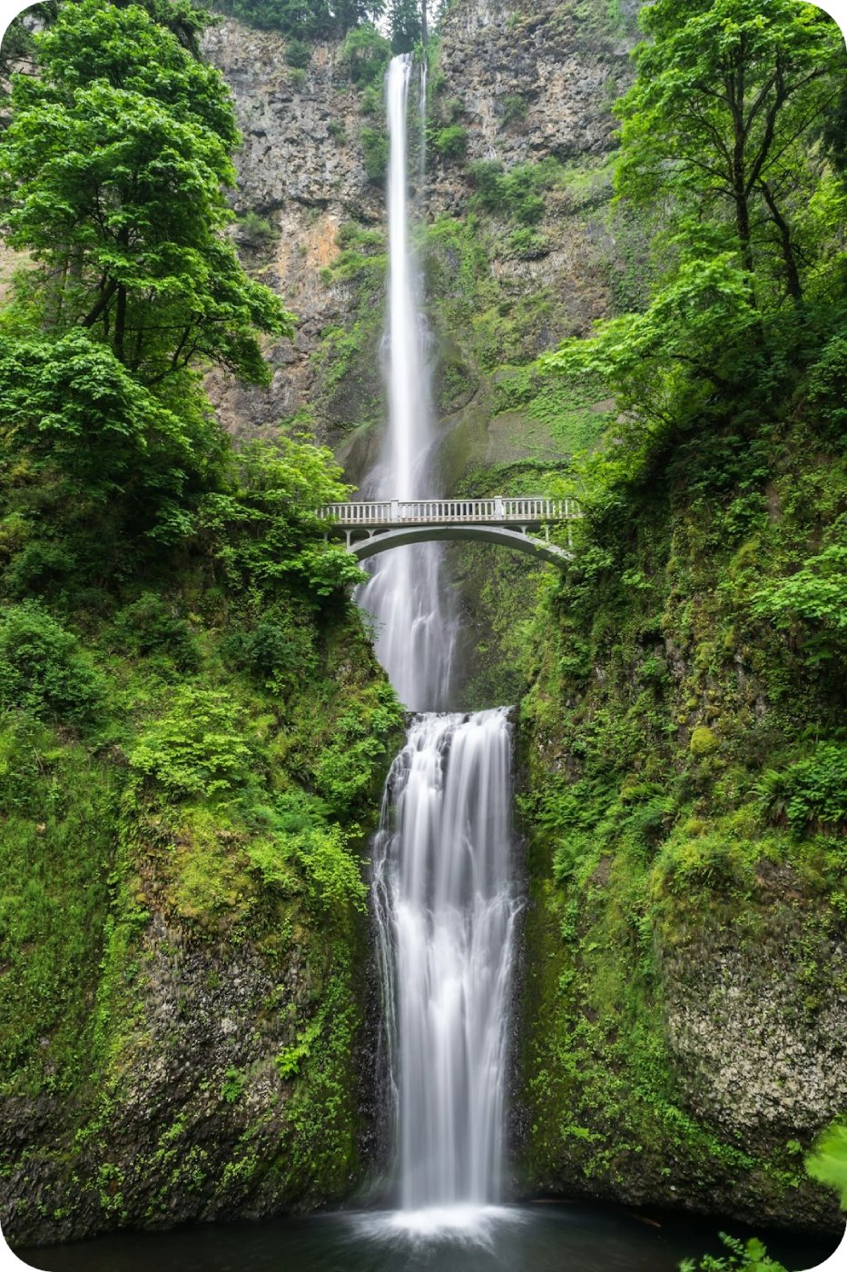 A tall two-tiered waterfall plunging down a mossy cliff, with a stone footbridge spanning the gorge between the upper and lower falls.
