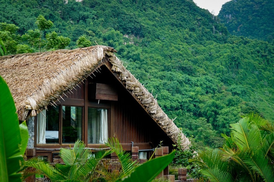 Wooden bungalow with a thatched palm roof and large windows, surrounded by tropical plants against a lush forested mountainside.