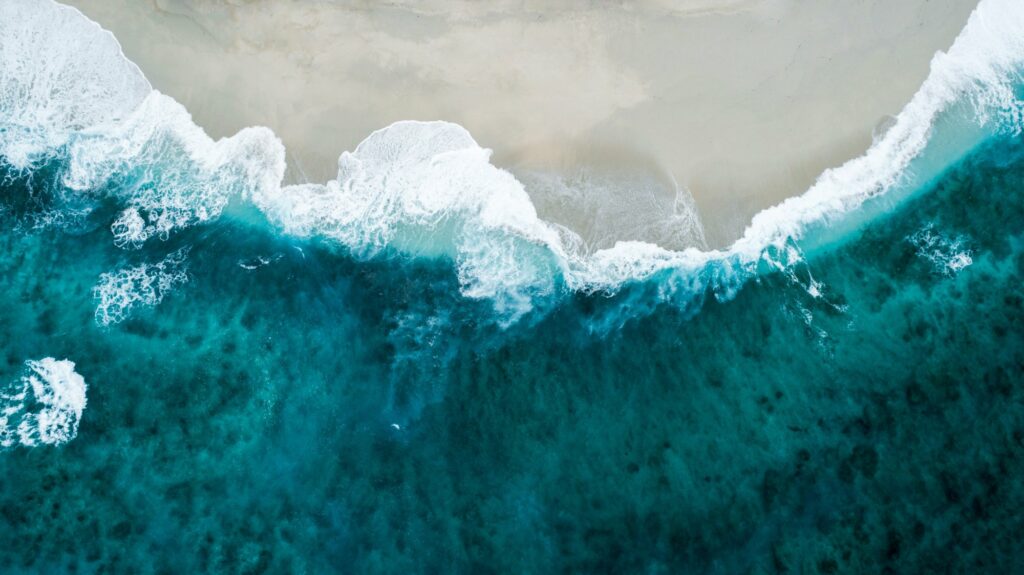 Aerial view of turquoise ocean waves breaking in white foam along a curved sandy beach.