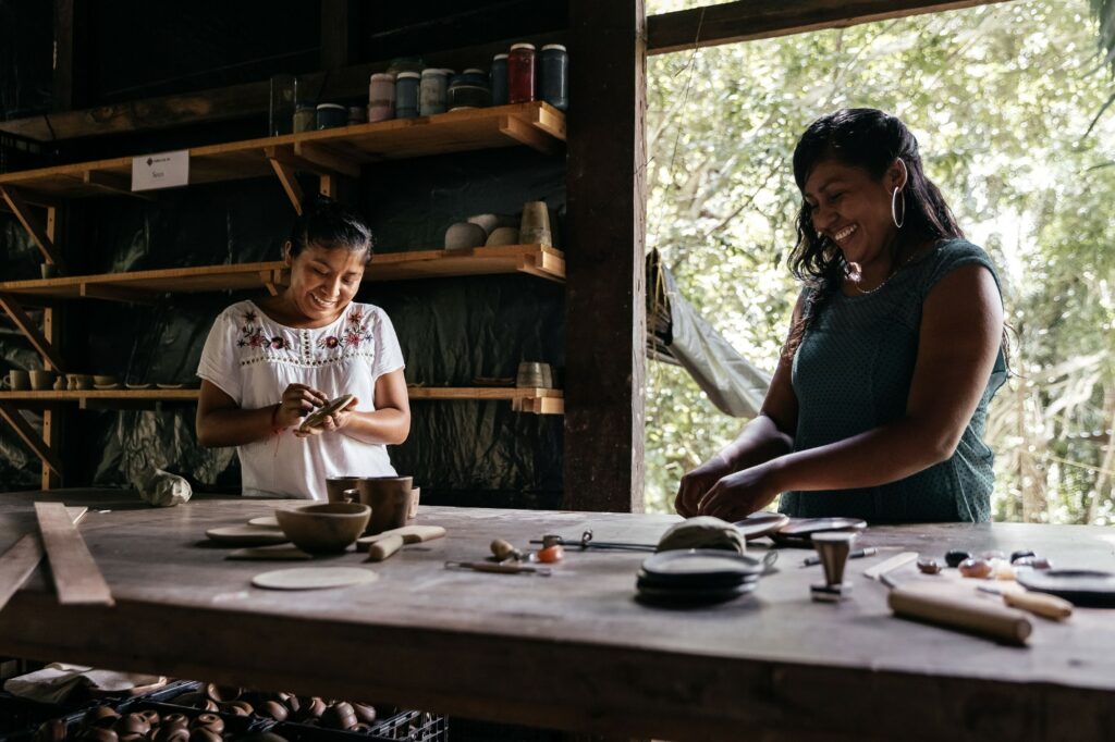 Two smiling women shaping clay pieces at a wooden workbench in an open-air pottery studio, with shelves of jars and ceramics behind them and jungle foliage visible outside.