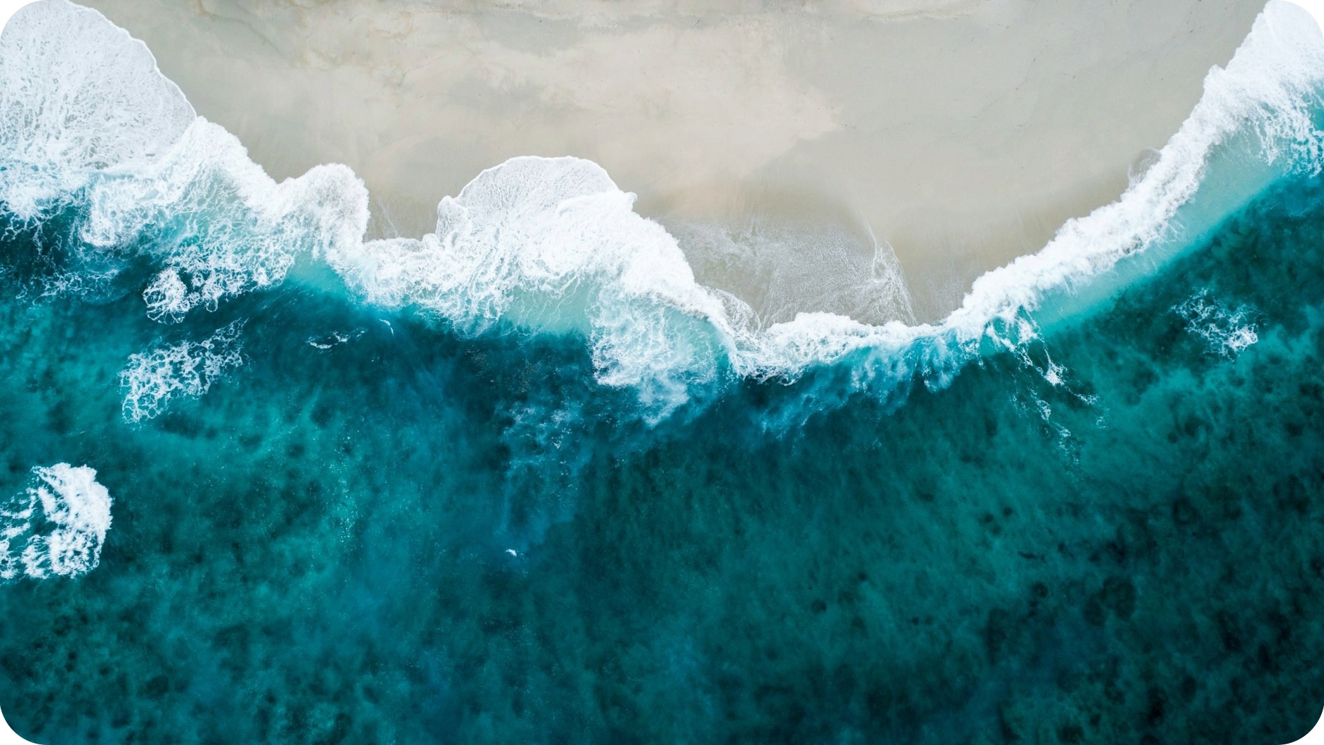 Aerial view of turquoise ocean waves breaking in white foam along a curved, sandy beach.