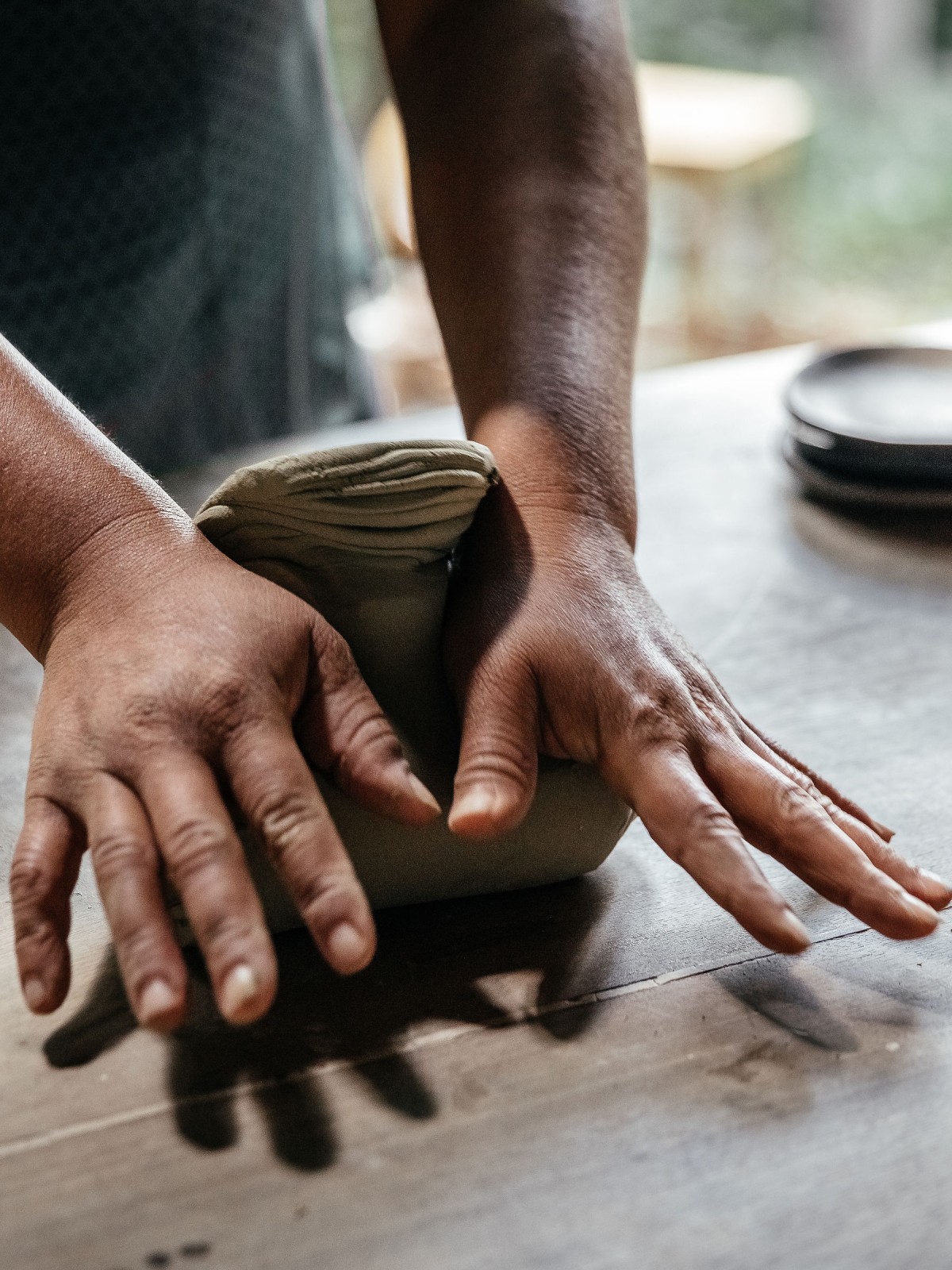 Close-up of a potter's hands kneading a block of clay on a wooden table, with dark ceramic plates blurred in the background.